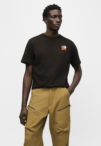 Νεαρός άνδρας που φοράει μαύρο μπλουζάκι The North Face και μουσταρδί παντελόνι με τσέπες με φερμουάρ, στέκεται με το ένα χέρι στην τσέπη.