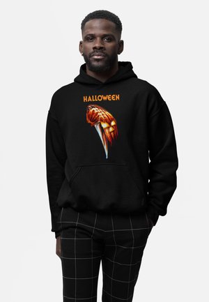 Mężczyzna noszący czarną bluzę Halloween z kapturkiem z motywem noża i twarzy dyni, połączoną z czarnymi spodniami z białymi liniami siatki.
