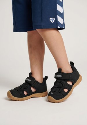 Kind trägt schwarze und braune geschlossene Sandalen und marineblaue Shorts mit weißen Zickzackstreifen auf hellem Hintergrund.