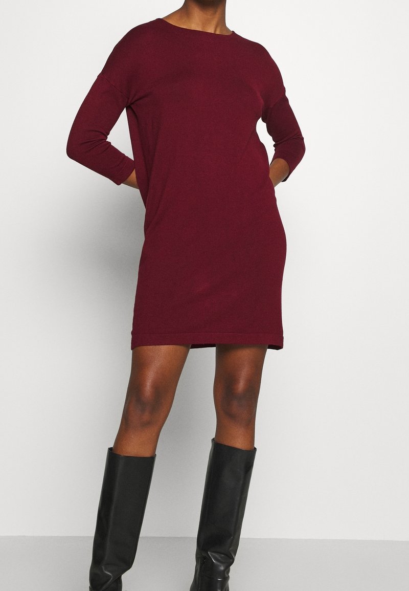 Femme portant une robe-pull bordeaux à manches longues associée à des bottes noires jusqu'aux genoux, debout devant un fond clair uni.