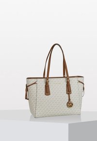 Sac fourre-tout beige Michael Kors avec des poignées en cuir marron et un charm en forme de logo, posé sur une surface blanche contre un fond uni.