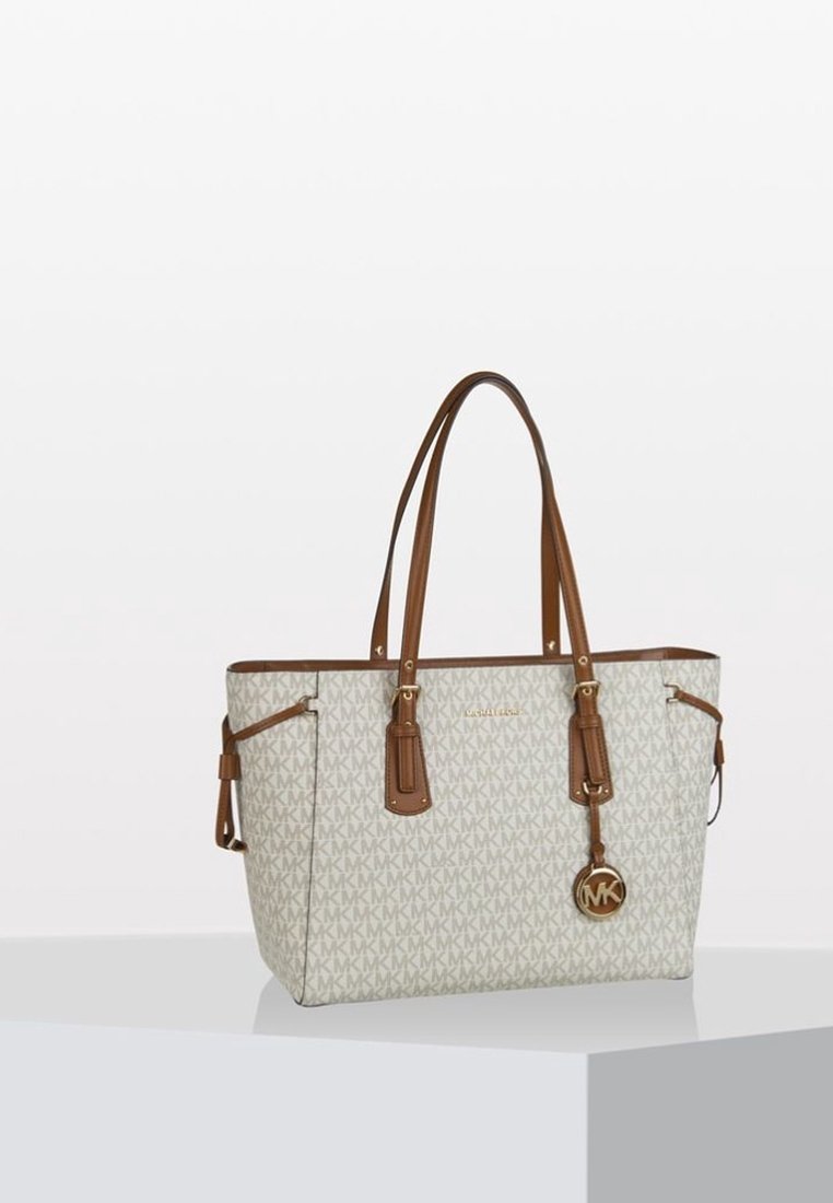 Sac fourre-tout beige Michael Kors avec des poignées en cuir marron et un charm en forme de logo, posé sur une surface blanche contre un fond uni.