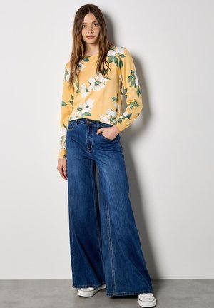 Żółty top z długim rękawem w biały kwiatowy wzór, zestawiony z szerokimi niebieskimi jeansami i białymi trampkami. Modelka stoi na tle neutralnego tła.