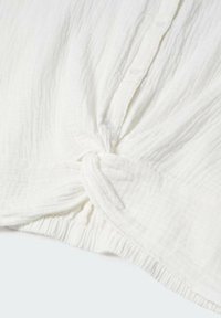 Biały, teksturowany top wykonany z tkaniny, z węzłem na dole i guzikami. Materiał ma pomarszczony wzór, co dodaje mu głębi.