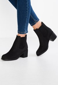 Botas de tobillo negras hechas de un material similar al ante, con paneles laterales elásticos, un tacón ancho y un diseño de punta redondeada y suave.