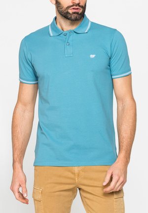 Uomo che indossa una polo blu a maniche corte con rifiniture bianche e pantaloni cargo color senape, in piedi davanti a uno sfondo semplice.