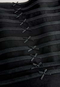 Schwarze Panties mit Spitze, in Reihen angeordnet, die vorne kleine Satin-Schleifen haben. Der Stoff ist glatt und hat eine weiche Textur.