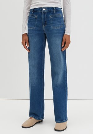 Person iført løstsiddende blå cowboybukser med frontlommer, en hvid langærmet top og beige slip-on sko.