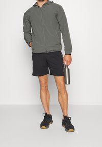 Chaqueta ligera con capucha en verde oliva, combinada con pantalones cortos deportivos negros. Zapatillas negras y una botella de agua plateada completan el conjunto.