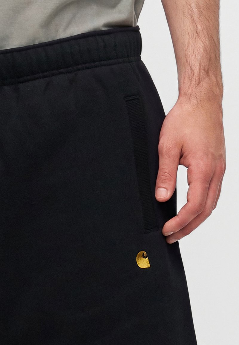 Nahaufnahme einer Hand, die in der Nähe der Tasche schwarzer Jogginghosen mit einem kleinen gelben Logo am Oberschenkel ruht.