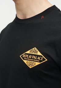 Μαύρο T-shirt με στρογγυλή λαιμόκοψη και κίτρινο διαμαντένιο λογότυπο που γράφει "Miami Replay Palace 81st Marcoari Avenue Florida" στο αριστερό στήθος.