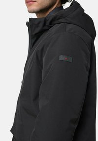 Zwarte jas met capuchon, gemaakt van gladde stof. Voorzien van een rits en een merkpatch op de mouw. Strakke, getailleerde ontwerp.