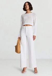 Pantaloni bianchi a vita alta e gamba larga abbinati a un top corto bianco lavorato a maglia con motivo a diamante aperto. Tiene in mano una borsa color cuoio.