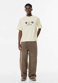 Hellgelbes T-Shirt mit Grafiken von Sonne und Erde; lockere Passform; kombiniert mit übergroßen braunen Hosen und neutralen Schuhen.