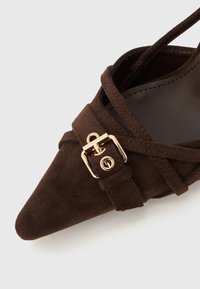 Rjava semiš čevelj z ostrim koničastim prstom in prekrižanimi trakovi; ima detajl z zaponko v zlatem tonu. Notranjost je gladka z rahlim sijajem.