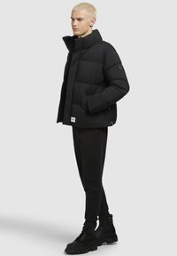 Schwarze Pufferjacke mit hohem Kragen, gestepptem Design, Reißverschlusstaschen und einem Logo-Patch am Saum, kombiniert mit schwarzen Sporthosen und Stiefeln.