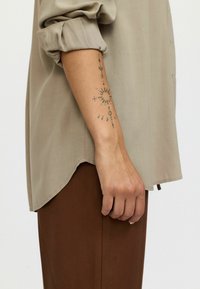 Beige skjorte med knapper og oprullede ærmer, med en buet kant. Brune stofbukser synlige, med et dekorativt tatovering på underarmen.