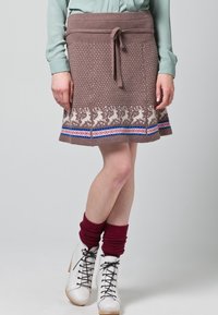 Jupe en tricot marron avec motif de renne, taille à cordon, et accents colorés en bleu et rouge sur l'ourlet. Portée avec des chaussures blanches et des jambières rouges.