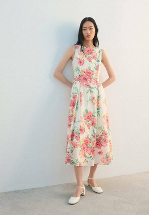 Frau steht vor einer weißen Wand und trägt ein ärmelloses weißes Midikleid mit pink-grünem Blumenmuster und weiße, niedrig-absatzige Schuhe.