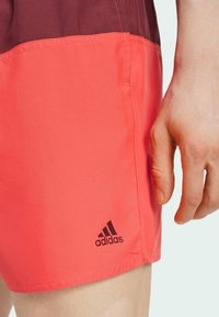 Pantaloni Adidas, di un vibrante colore corallo con una parte superiore bordeaux a contrasto. Presentano una texture liscia e un piccolo logo nero sul lato.