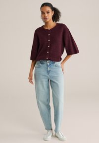 Tumši sarkans adīts džemperis ar īsām piedurknēm un lielām pogām, kopā ar gaiši ziliem vaļējiem džinsiem un baltām sporta kurpēm uz neitrāla fona.