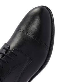 Zwarte leren nette schoen met gesloten vetersluiting, kapteen en decoratieve perforaties op een witte achtergrond.