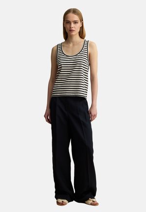 Femme portant un haut sans manches rayé noir et blanc, un pantalon noir large et des sandales beiges ouvertes à l'avant, debout devant un fond uni.