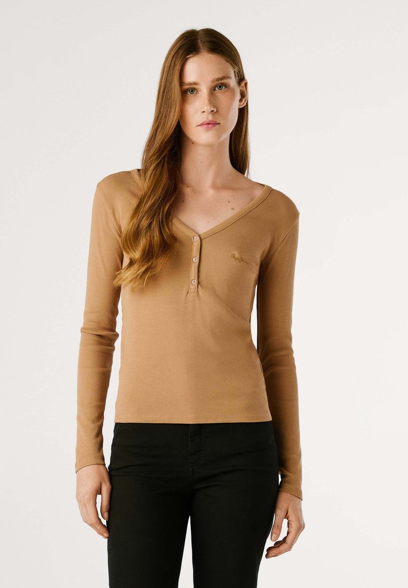 Langsleeve geribbelde top in een tan kleur met een V-halslijn, voorzien van drie knopen aan de voorkant en een subtiel logo aan de linkerkant.