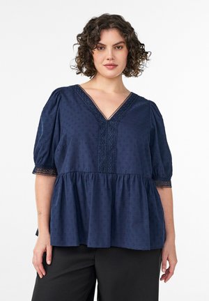 Žena s kučeravými vlasmi, ktorá nosí námornícky modrú blúzku s výstrihom do V, čipkovým lemom a nadýchanými rukávmi, spojenú s čiernymi nohavicami, stojaca na bielom pozadí.