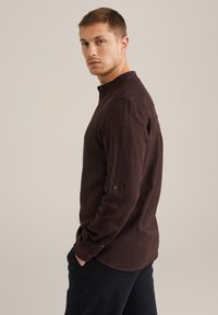 Cămașă cu mânecă lungă, de culoare maro închis, cu guler cu nasturi și manșete cu un singur nasture, fabricată dintr-un material texturat, asortată cu pantaloni negri.