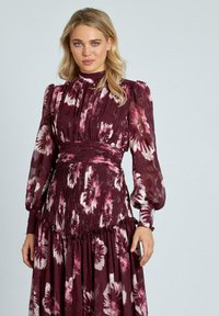 Vestido largo floral en burdeos con estampados en rosa y blanco. Presenta pliegues, un lazo en la cintura y mangas abullonadas. Está hecho de una tela ligera.