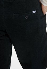 Schwarze Cordhosen mit ribbed Textur, vorderer Tasche und Knopfverschluss. Mit einem kleinen Etikett auf der Tasche.
