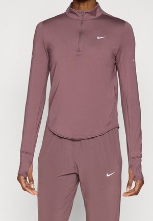 Personne portant un haut zippé mauve à manches longues et un pantalon assorti avec des logos blancs Nike sur la poitrine et la cuisse.