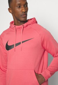 Pink hoodie with a black Nike logo, made from soft fabric. Features a front pocket, adjustable drawstring hood, and raglan sleeves.