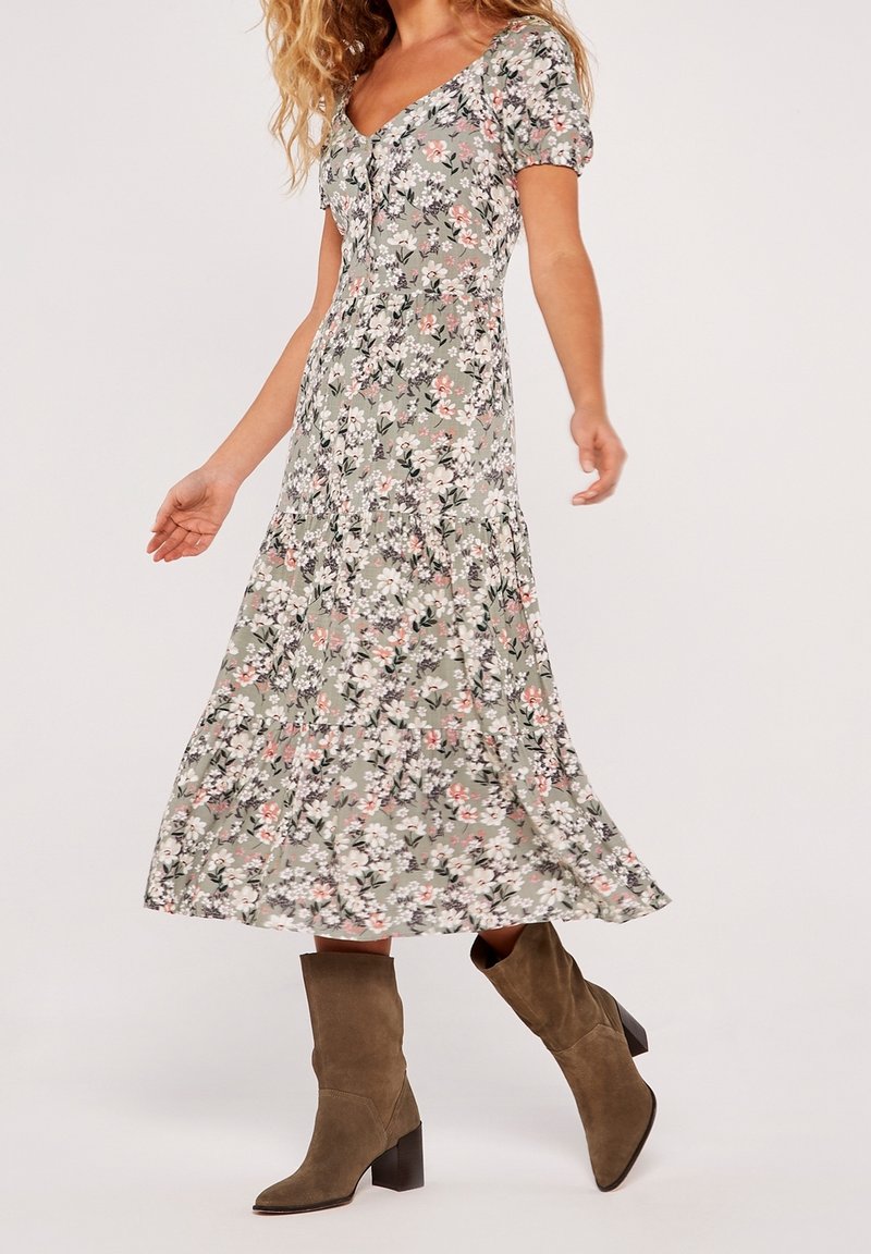 Femme portant une robe midi grise à fleurs avec manches bouffantes et des bottes à talons mi-mollet en daim marron, marchant devant un fond uni.