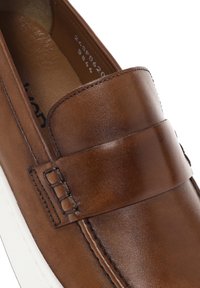 Mocasines de cuero marrón con una banda ancha en la parte frontal, costuras en contraste y una suela de goma blanca. Textura suave, forma redondeada.