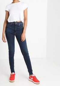 Blå smala jeans med hög midja, tillsammans med en vit kort ribbad t-shirt. Modellen har på sig ljusröda sneakers med vit sula.