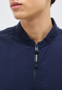 Κοντινό πλάνο ενός ατόμου που φοράει ένα μπλε σκούρο μπουφάν με ορατό φερμουάρ μπροστά, με την ετικέτα "ECOALF".