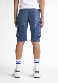 Modré denimové cargo šortky s několika kapsami, délka po kolena a vybledlý vzhled. V kombinaci s bílými ponožkami s modrými pruhy a botami s zelenými akcenty.