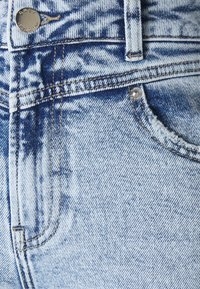 Tela de mezclilla azul claro con un patrón desvaído, que presenta un botón plateado y costuras de contraste a lo largo de las costuras y bolsillos.