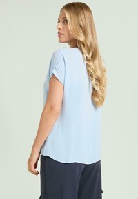 Lyseblå kortærmet top lavet af blødt stof med afrundet kant og afslappet pasform. Modellen har bølget hår og bærer mørke bukser.