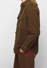 Camisa de franela marrón con botones, dos bolsillos en el pecho, textura acanalada y cuello, llevada sobre una camiseta roja y combinada con pantalones de franela marrón.