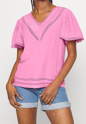 Kvinde iført en lyserød kortærmet bluse med blonder og opbukket blå denimshorts, stående med hænderne hvilende nær hofterne.