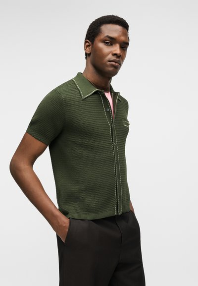 Άνδρας που φοράει κοντομάνικο πουκάμισο σκοτεινού πράσινου χρώματος με υφή και λευκές ραφές, μαύρο παντελόνι, στέκεται με τα χέρια στις τσέπες σε λευκό φόντο.