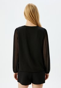 Blusa negra con mangas abullonadas y transparentes, y un cuello redondo. El material es suave con una textura fluida. Combinada con pantalones cortos negros.