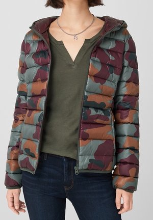 Femme portant une veste matelassée à motif camouflage sur un t-shirt vert olive à col en V et un jean bleu foncé, avec un collier à chaîne en argent.