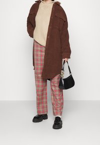 Brun fleecejacka över en beige stickad hoodie, matchad med röd och beige rutiga byxor samt svarta klumpiga skor; håller en svart väska med krokodilprägling.