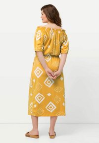 Keltainen, olkapäiden yltä laskeutuva mekko, jossa on valkoisia geometrisiä kuvioita, joustava kaula-aukko, lyhyet puffihihat ja istuva vyötärö. Tasainen kangaspinta.