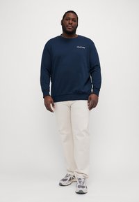 Marineblaues Sweatshirt mit einem "Club1990"-Logo, kombiniert mit beigen Hosen und grauen Turnschuhen, hat eine lockere Passform und lange Ärmel.