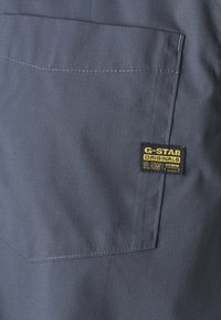 Sötétkék szövet, sima textúrával, látható zsebbel és fekete címkével, amelyen sárga betűkkel olvasható a "G-Star Originals".